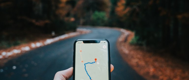 Phone in hand showing a route from A to B. Background shows a twisty road.